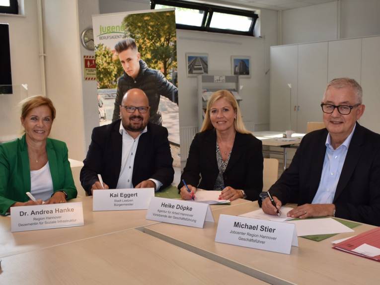 Zwei Männer und zwei Frauen sitzen an Konferenztischen und unterschreiben Dokumente. Vor Ihnen sind Namensschilder.