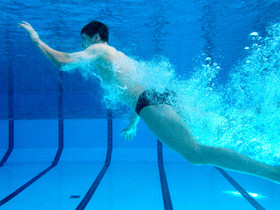 Ein Mann in schwarzer Badehose taucht in einem Schwimmbecken. Die vielen Luftblasen hinter und unter ihm lassen vermuten, dass er gerade von einem Sprungturm in das Becken gesprungen ist und nun wieder in steiler Kurve auftaucht.