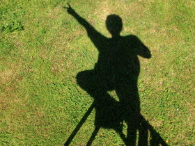 Auf einer hellgrünen Rasendläche zeichnet sich deutlich ein dunkler Schatten ab: Eine Person steht ganz oben auf einer Bockleiter. Mittelfinger und Zeigefinger bilden an einem ausgestreckten Arm die Victory-Geste.