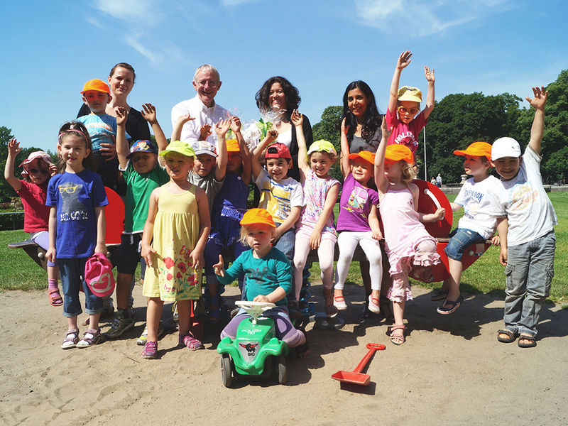 Kita-Kinder stehen an einer Wippe, im Hintergrund vier erwachsene Personen