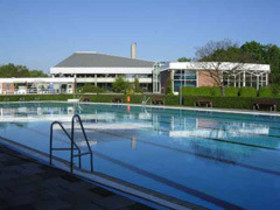 Das mit Wasser gefüllte Aussenbecken des Hallenfreibades Burgdorf ohne Badegäste bei strahlend blauem Himmel, im Hintergrund die Gebäude des Hallenbades, die sich im Wasser spiegeln
