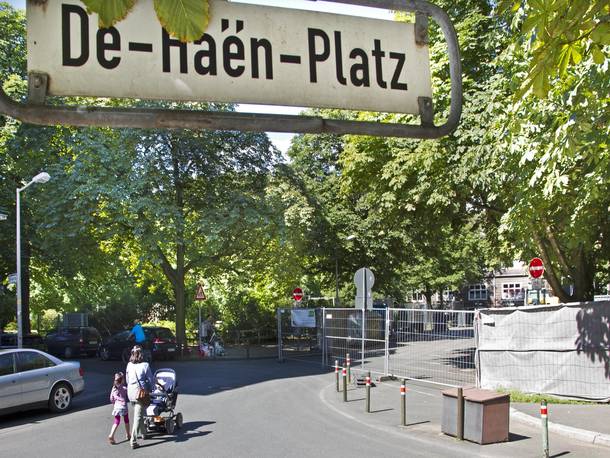 Straßenschild De Haen Platz, im Hintergrund eine Frau mit Kinderwagen und eine Baustellenabsperrung