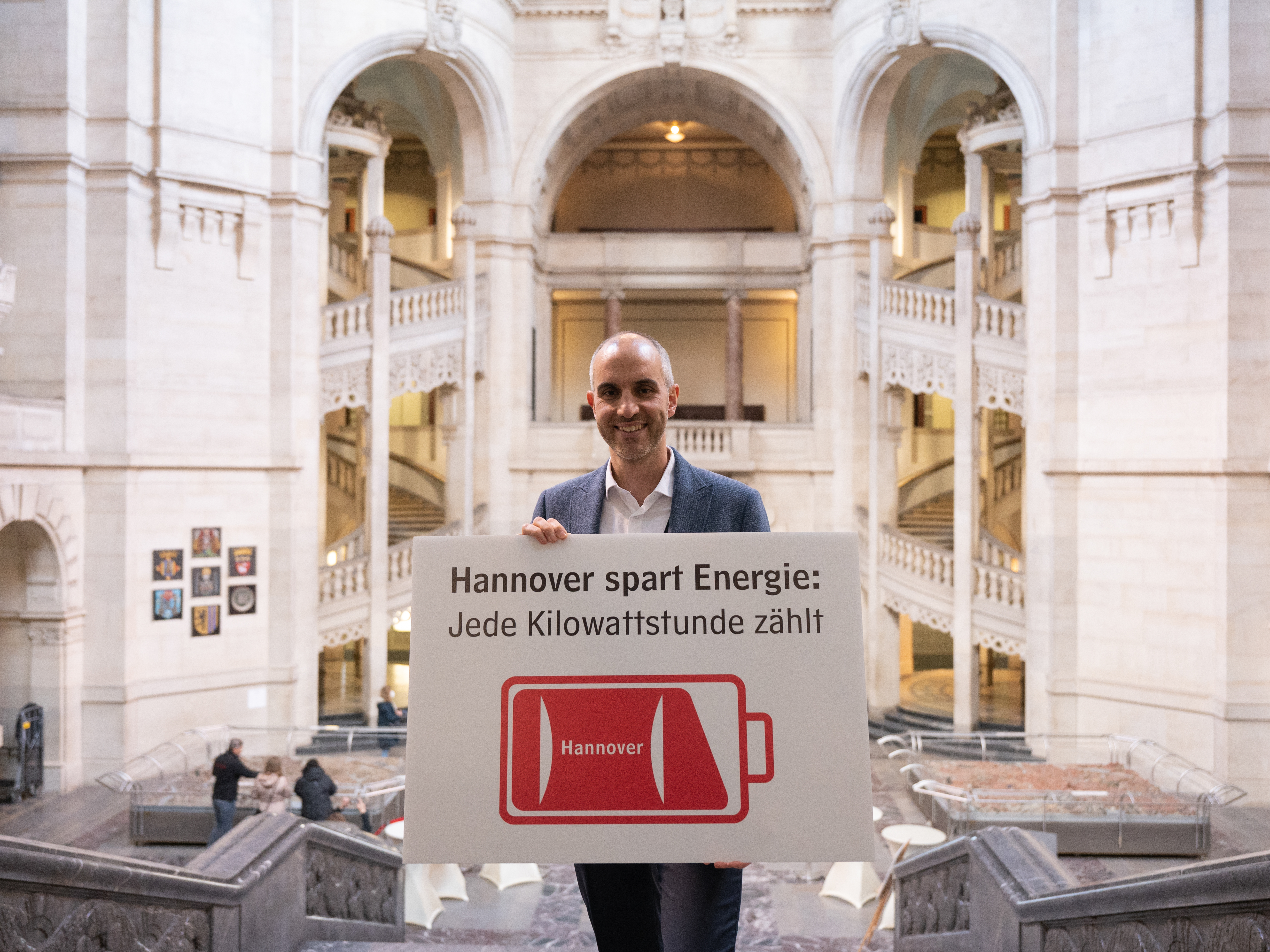 Ein Mann steht in der Rathaushalle und hält ein weiß-rotes Schild mit einem Batterielogo und der Aufschrift "Hannover spart Energie: Jede Kilowattstunde zählt".