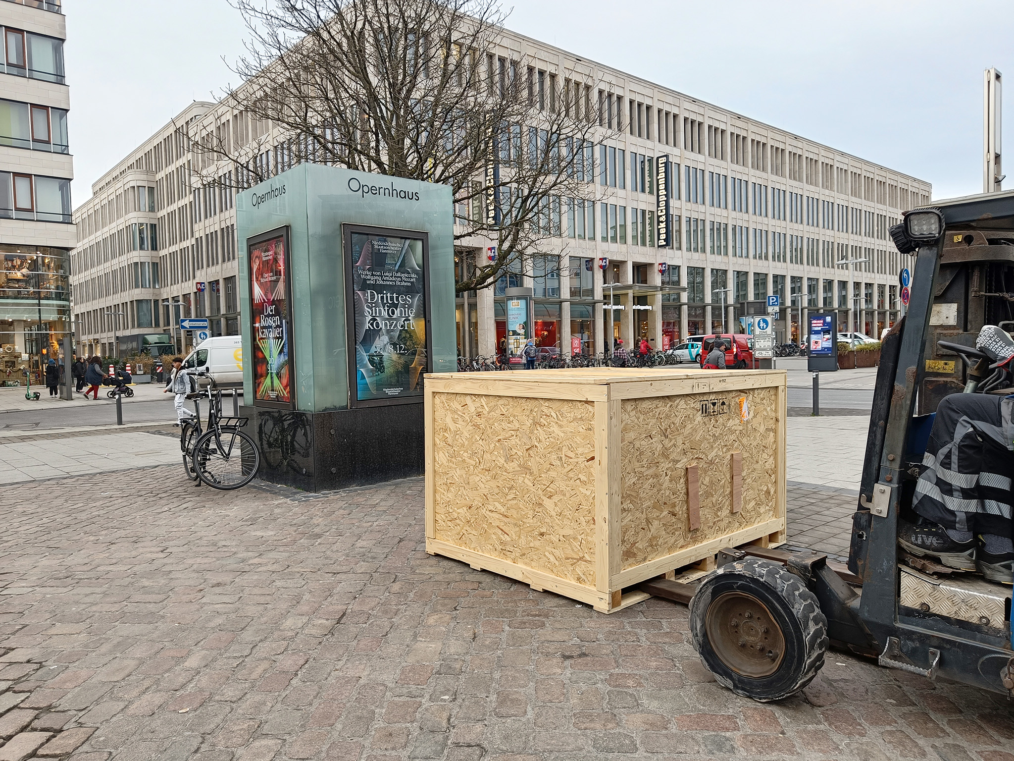 Auf einem Platz steht ein Gabelstapler, der eine Holzkiste auf den Boden abgestellt hat, davor ein Schaukasten mit der Aufschrift Opernhaus.