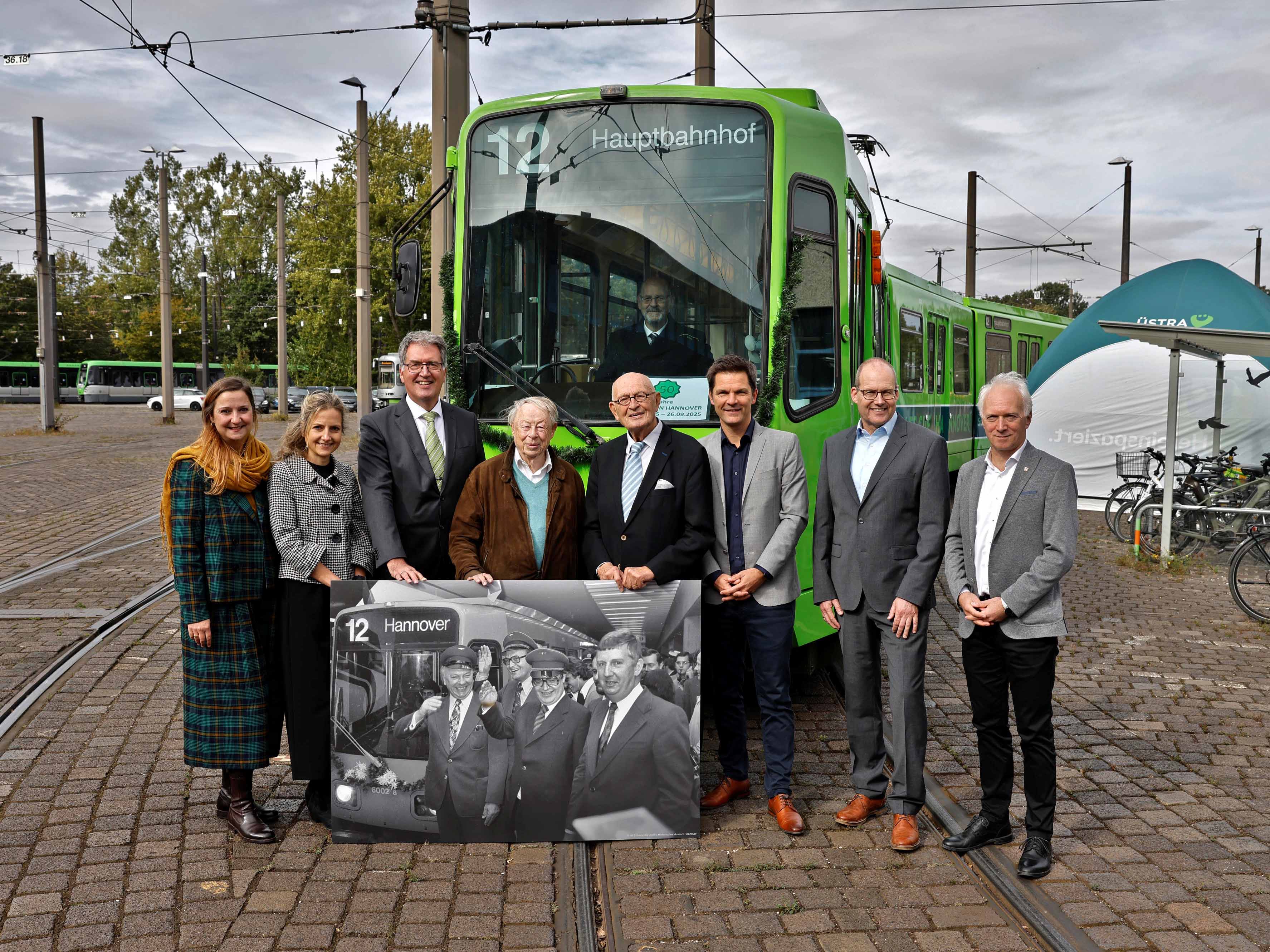 Eine Gruppe von Menschen steht vor einer Stadtbahn und präsentieren ein historisches Foto, auf dem die erste Stadtbahnfahrt von 1975 gezeit ist.