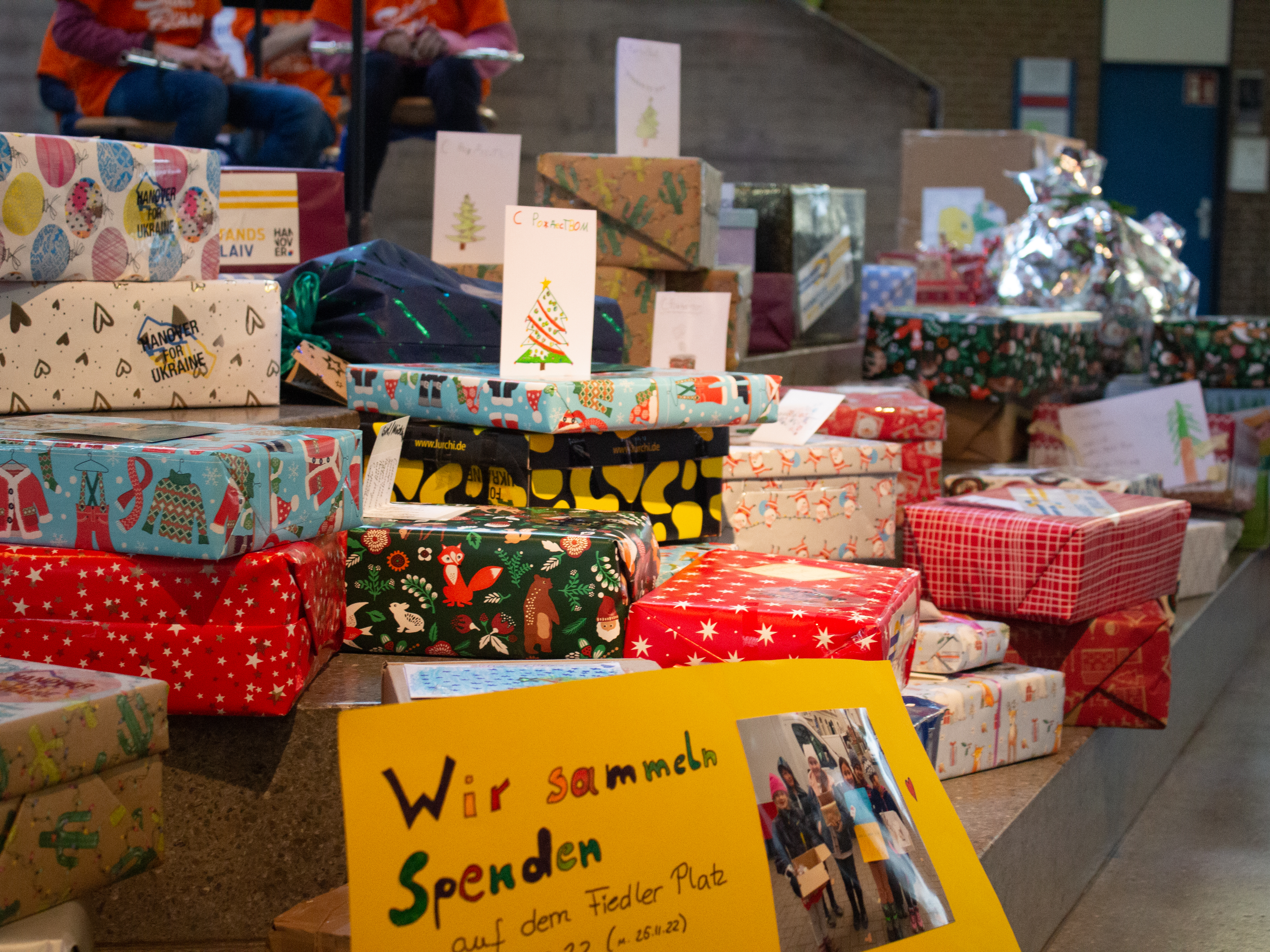 Bunt eingepackte Geschenke und im Vordergrund ein gelbes Schild mit der Aufschrift "Wir sammeln Spenden".