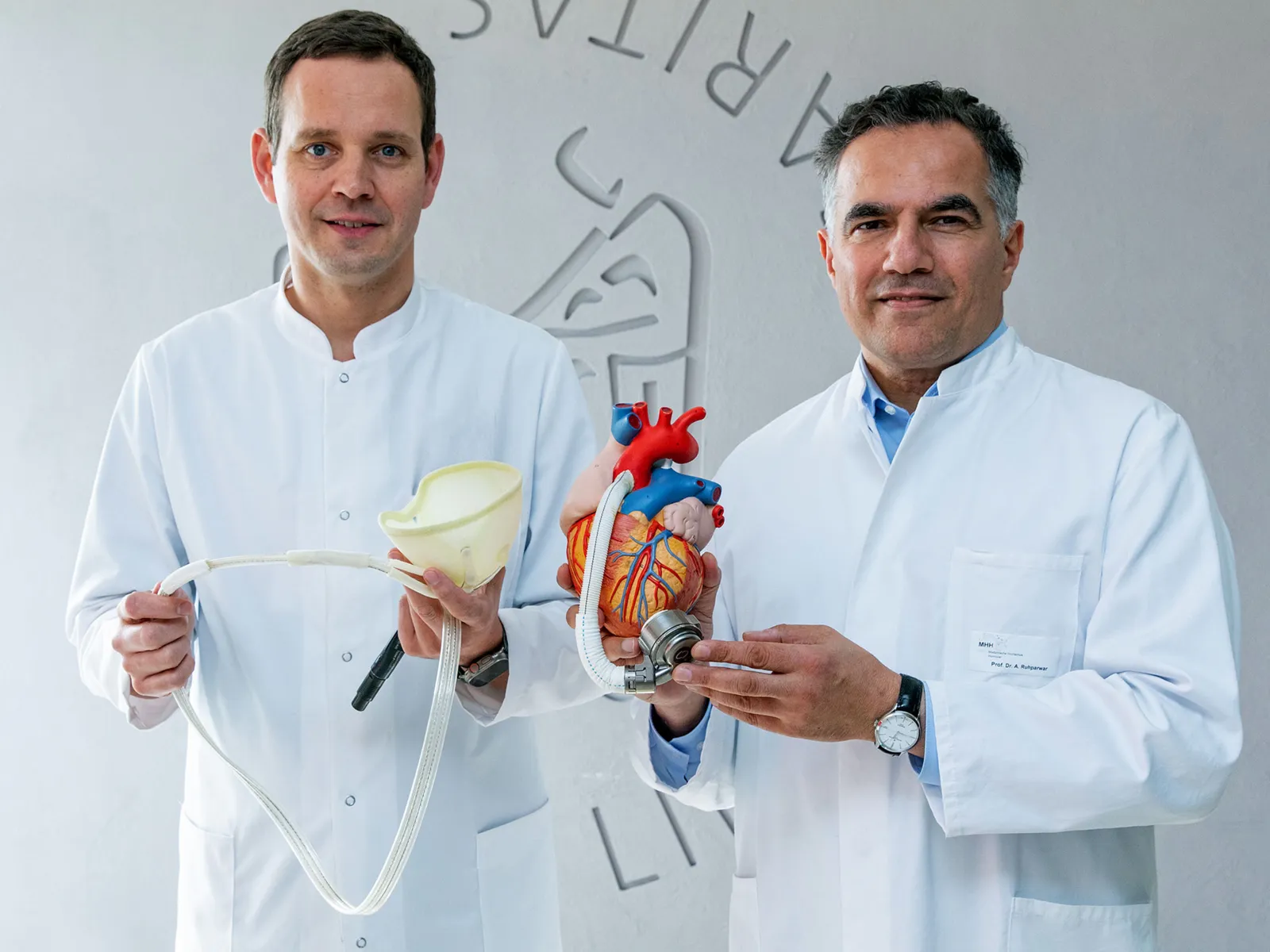 Zwei Männer in weißen Kitteln halten medizinische Geräte und ein Modell eines Herzen in den Händen.