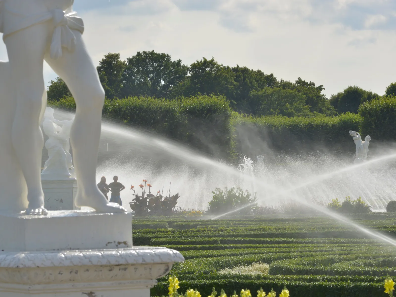 Vorn links die Beine einer weißen Skulptur auf einem Sockel, dahinter zwei überkreuzte Wasserstrahlen, aus denen es auf grüne Linien aus flachen Pflanzen regnet.