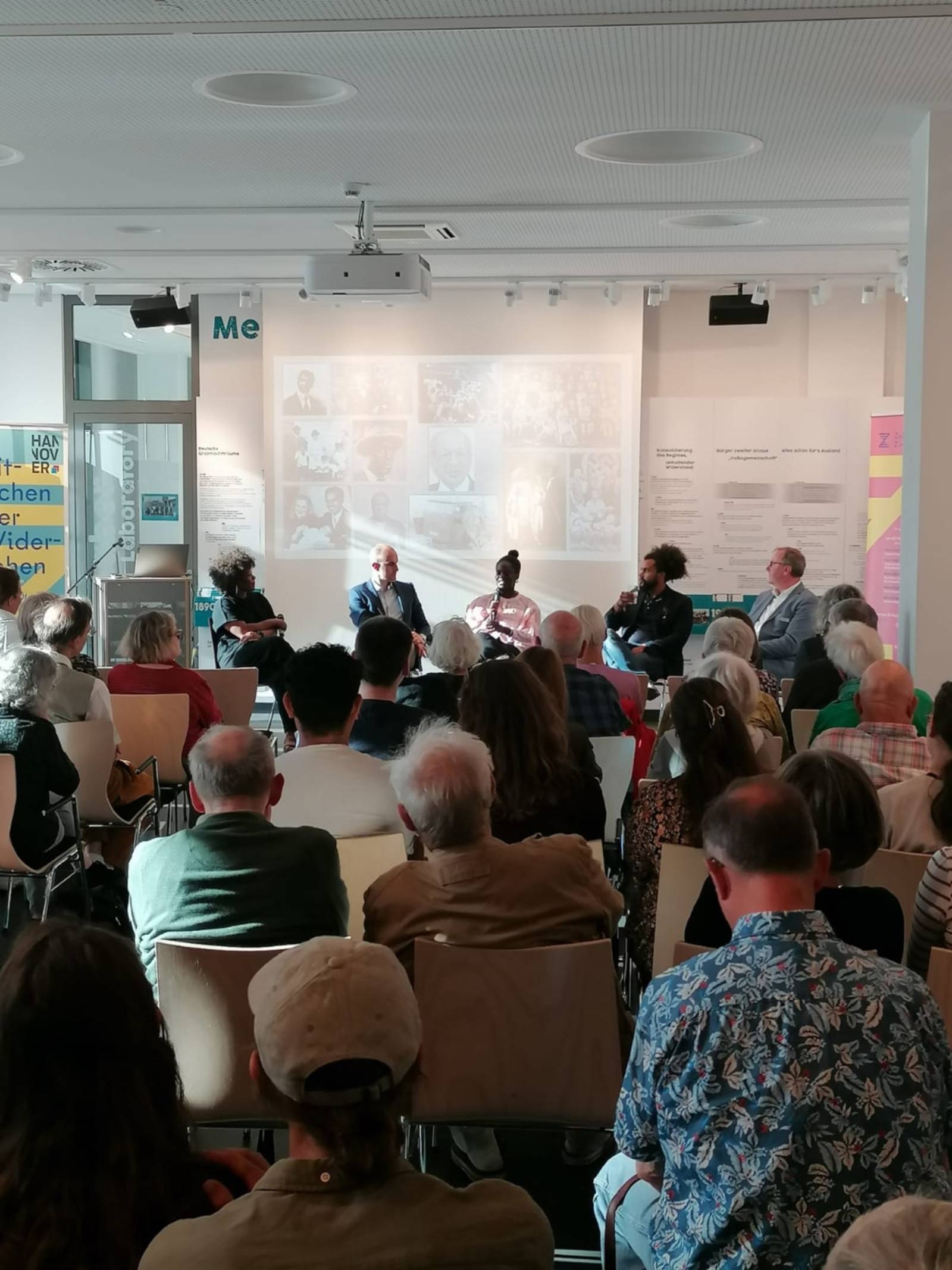 Eine Veranstaltung im ZeitZentrum Zivilcourage. Bild von hinten aus dem Publikum. Zu sehen sind gefüllte Reihen von Hinterköpfen, sie sind alle auf das Podiumsgespräch gerichtet. Im Podium sitzen fünf Personen, darunter Brenda Davina, Dr. Jens Binner und Oberbürgermeister Belilt Onay. Brenda Davina spricht in das Mikrofon.