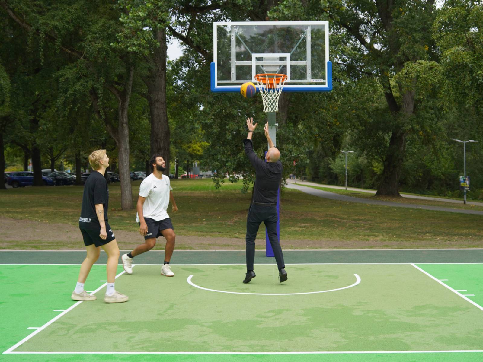 Zwei M&auml;nner in Sportkleidung, einer in schwarzer Hose  und schwarzem Hemd auf einem gr&uuml;nen Sportfeld. Der Mann im schwarzen Hemd springt vor dem Basketballkorb und wirft den Ball hinein.