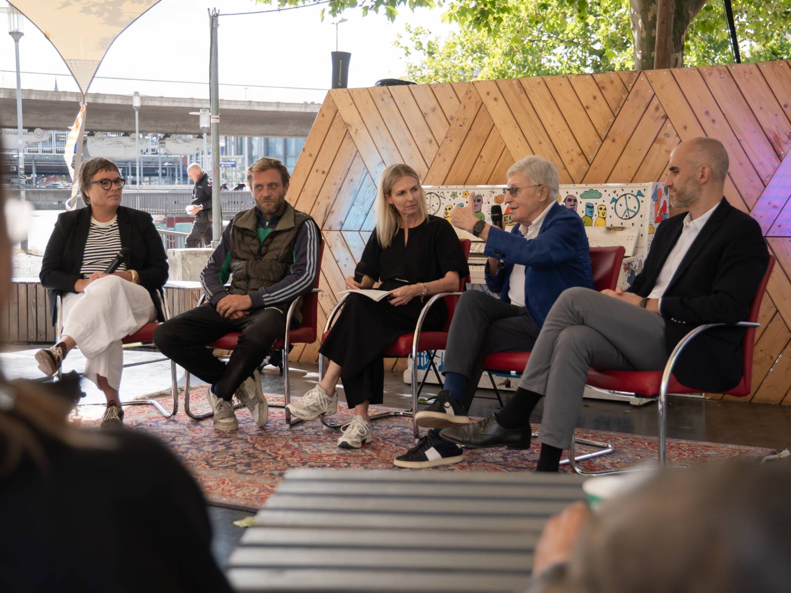 Vier Menschen sitzen auf Stühlen auf einer kleinen Bühne, ein Mann spricht in ein Mikrofon.