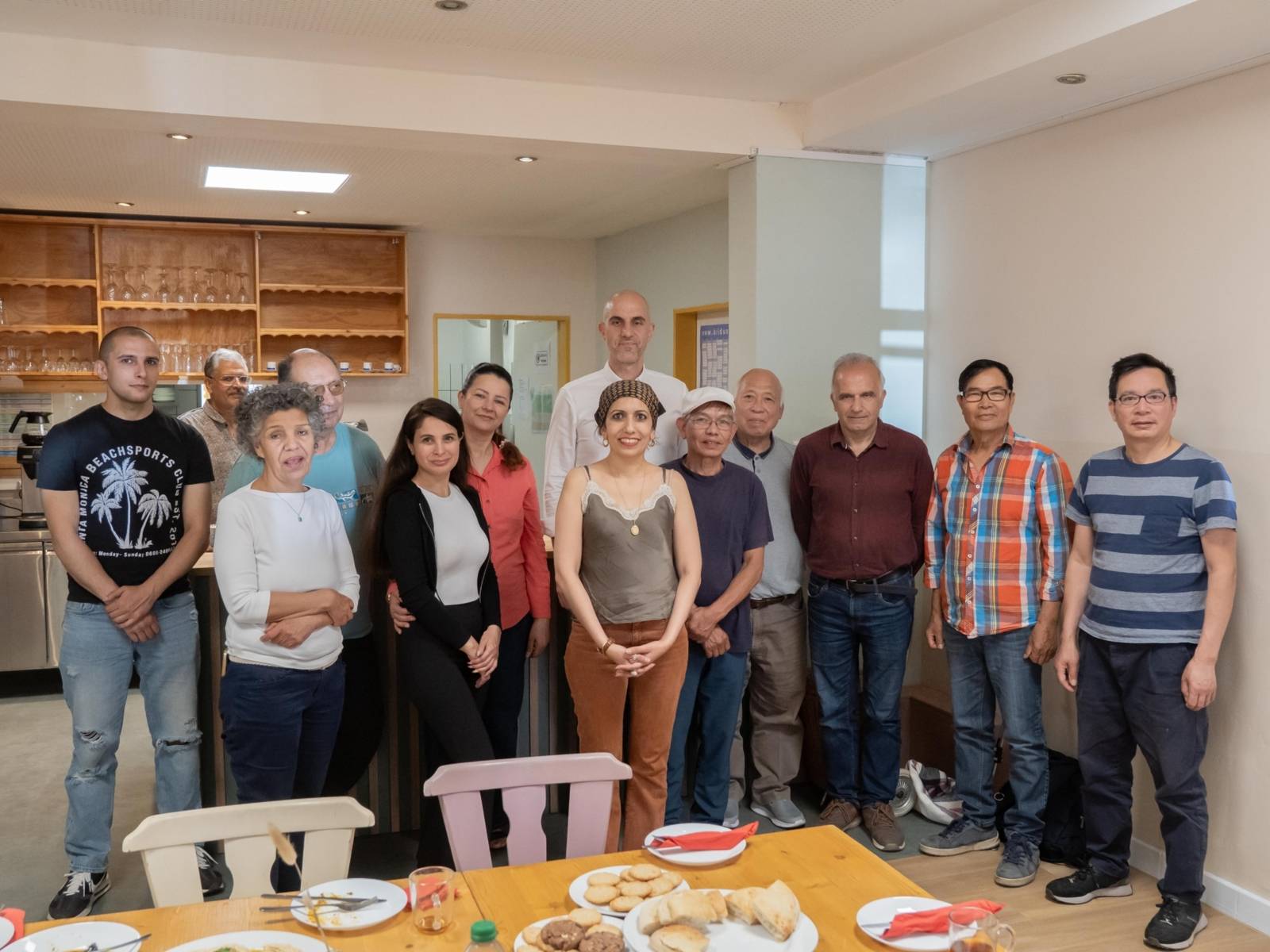Ein Gruppenbild. Im Vordergrund ein gedeckter Esstisch, in der Mitte eine Gruppe von 13 Personen mit Oberb&uuml;rgermeister Belit Onay in der Mitte. Im Hintergrund eine h&ouml;lzerne Bar und K&uuml;chenzeile.