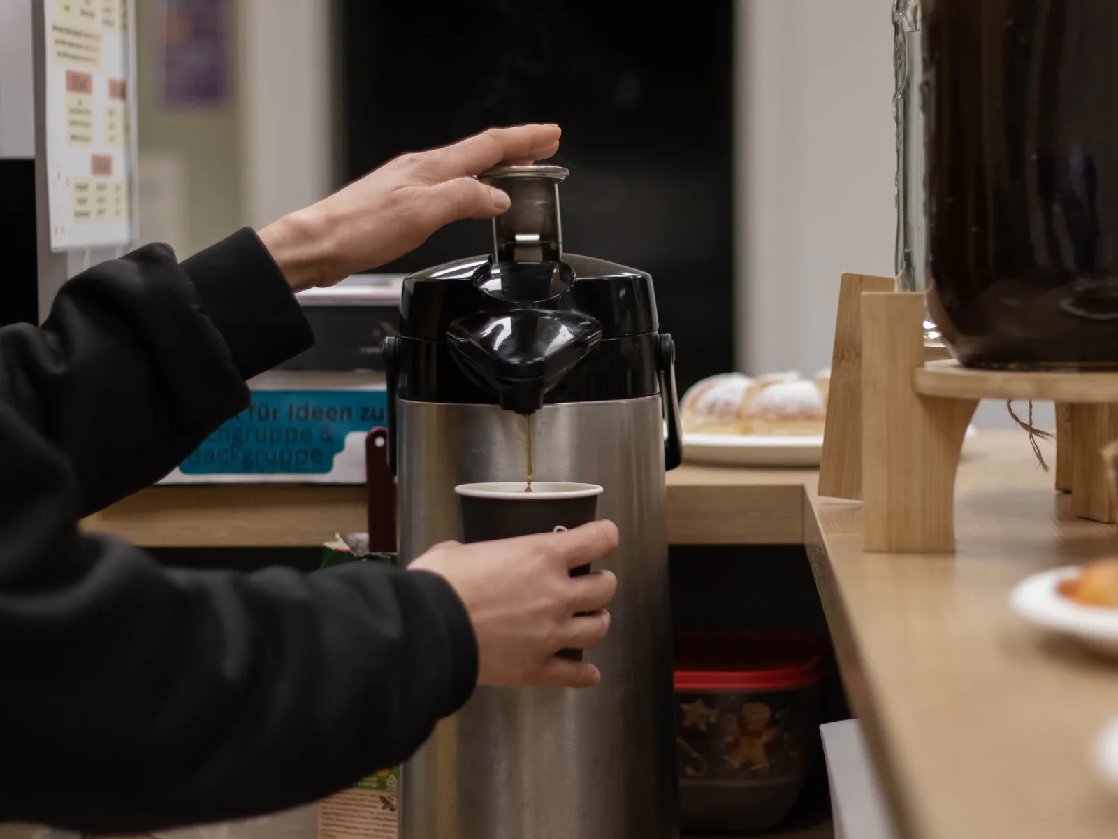 Eine Theke von der Seite in einem Raum, in der Mitte eine große Thermoskanne. Zwei Hände füllen per Knopfdruck eine dunkle Flüssigkeit in einen Becher.