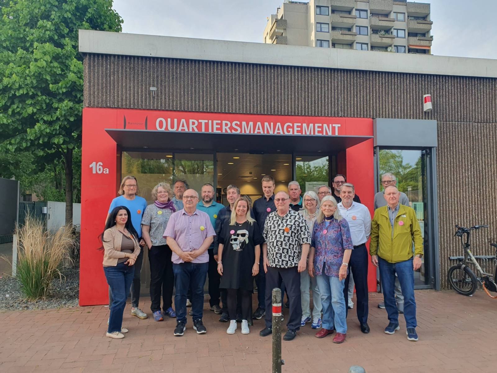 Eine Gruppe von Menschen steht vor einem Gebäude mit einem roten Rahmen um eine Doppeltür, auf dem Quartiersmanagement steht.
