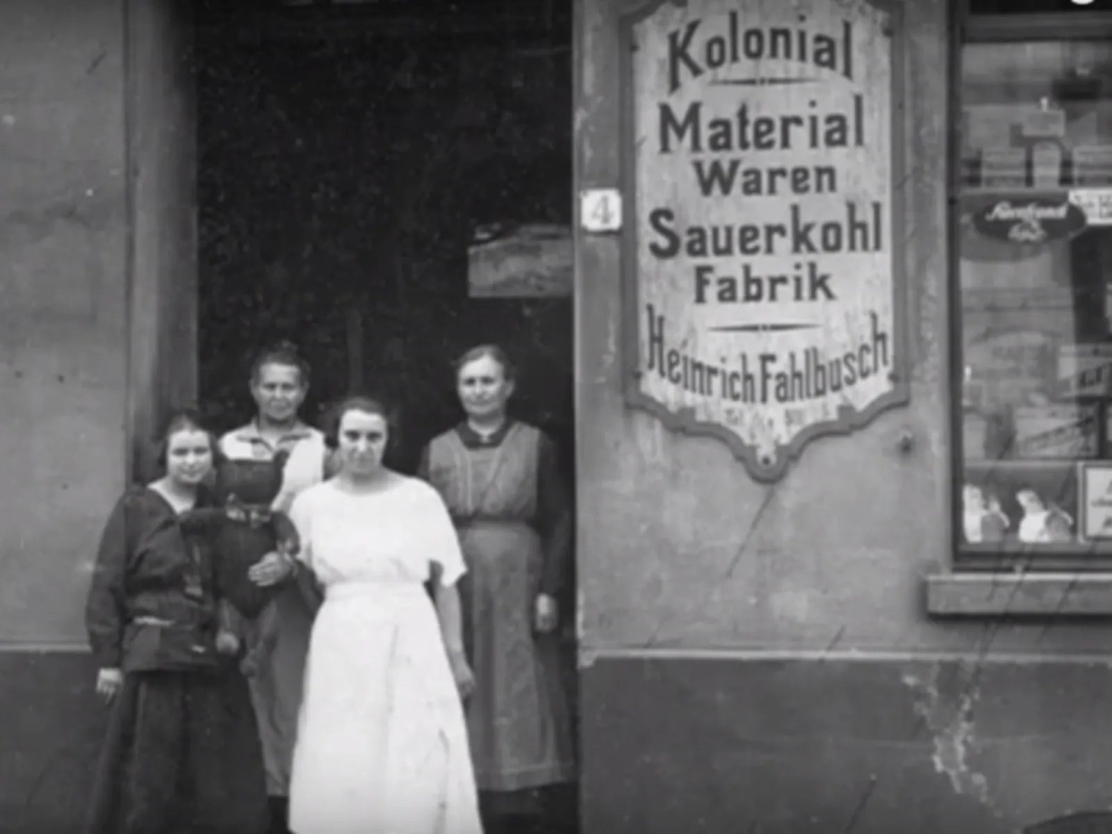 Ein altes Schwarz-Weiß-Foto: Vier Frauen in einem Hauseingang neben dem Schild "Kolonial Material Waren Sauerkohl Fabrik Heinrich Fahlbusch.