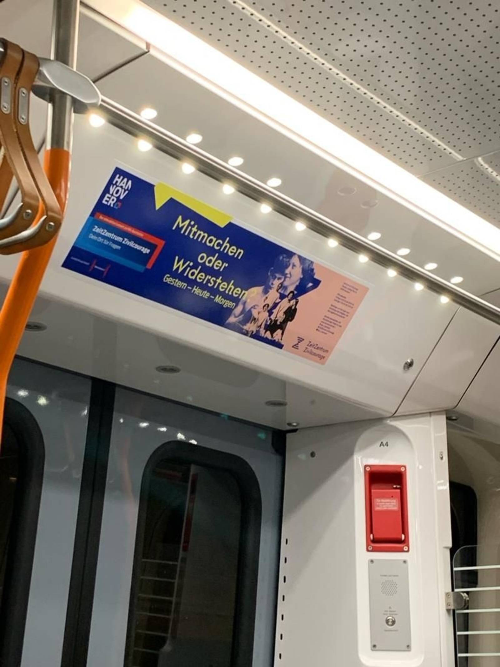 Zu sehen ist ein Banner über der Tür im Innenraum einer Straßenbahn. Der Banner besteht aus Werbung für das ZeitZentrum Zivilcourage. Groß zu sehen ist die Frage "Mitmachen oder Widerstehen?".