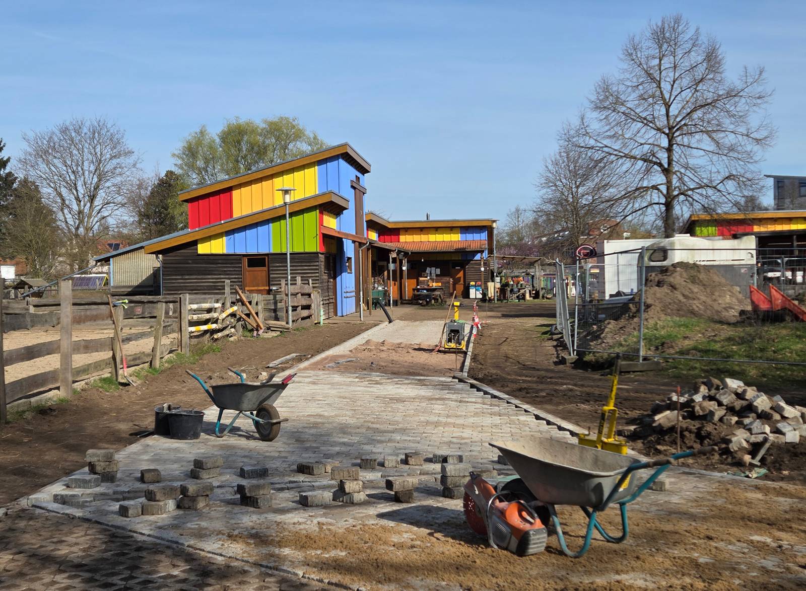 Zwei Schubkarren und Gehwegsteine vorn, dahinter ein teilweise fertiger Weg, rechts ein Bauzaun, links ein Holzgatter und daneben weiter hinten ein bunt gestreiftes Haus mit zwei Geschossen.