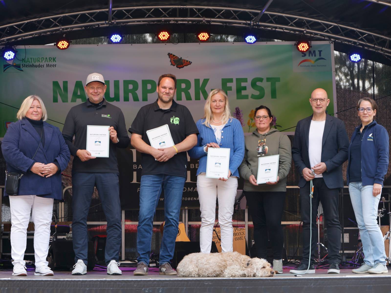 Sieben Personen stehen auf einer B&uuml;hne, vier der Personen halten eine Urkunde in den H&auml;nden. Mittig zu deren F&uuml;&szlig;en liegt ein Hund.