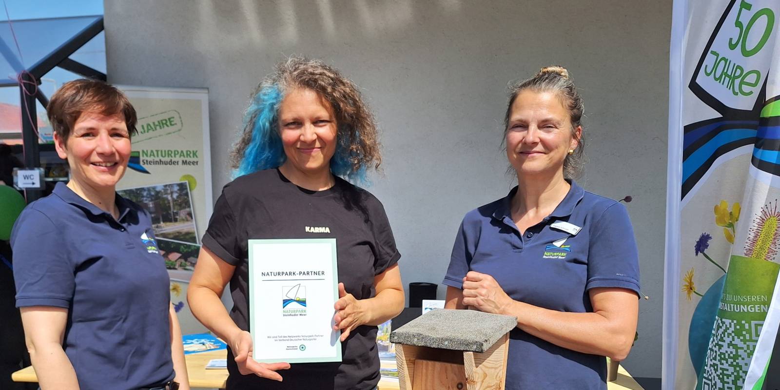 Drei Frauen stehen nebeneinander und schauen in die Kamera. Eine Frau hält dabei eine Urkunde, eine andere ein Vogelhäuschen.