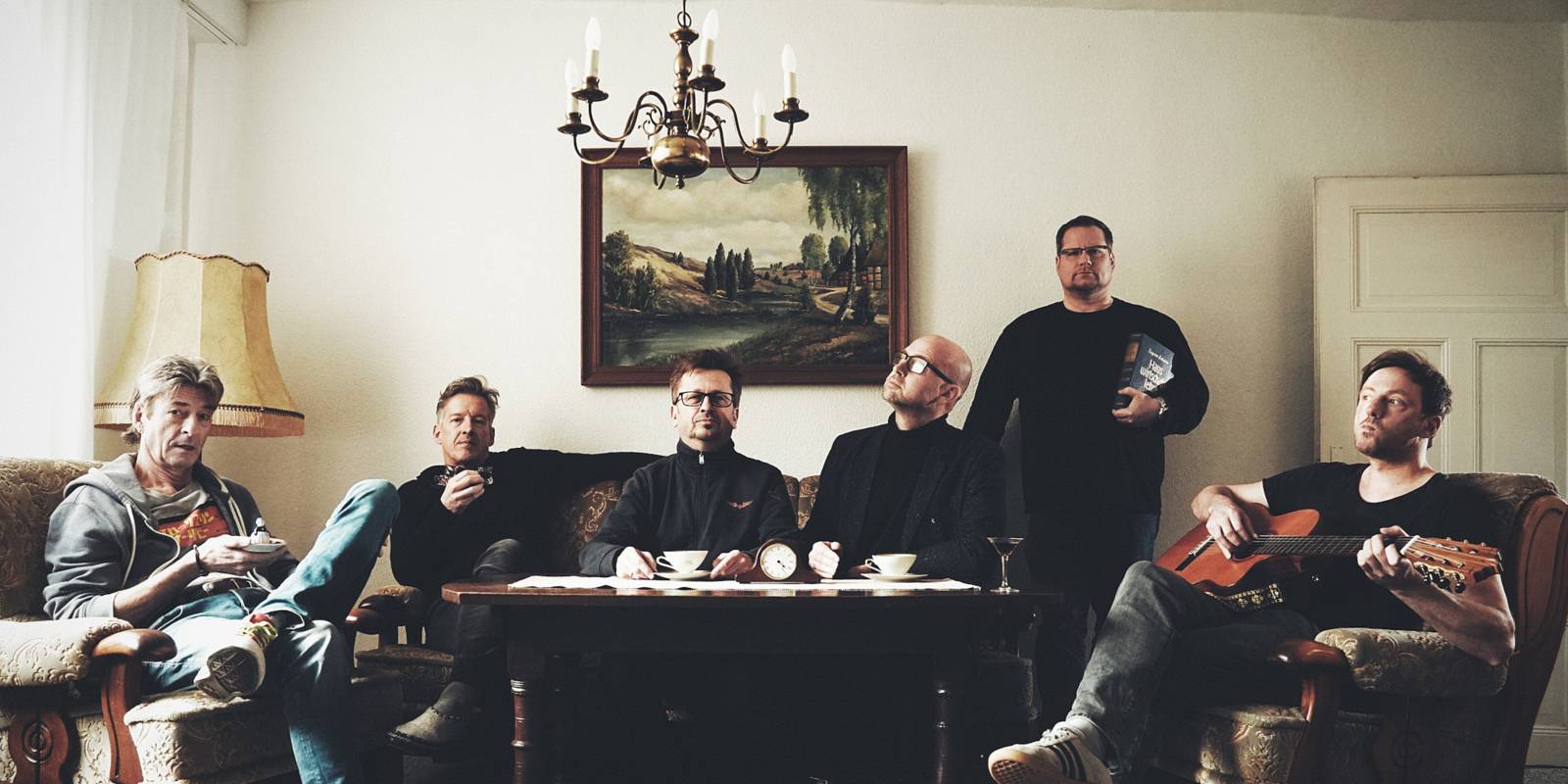 Sechs Herren sitzen bei Kaffee oder mit Instrument in rustikalem Wohnzimmerambiente – die Herberts vom Salon Herbert Royal