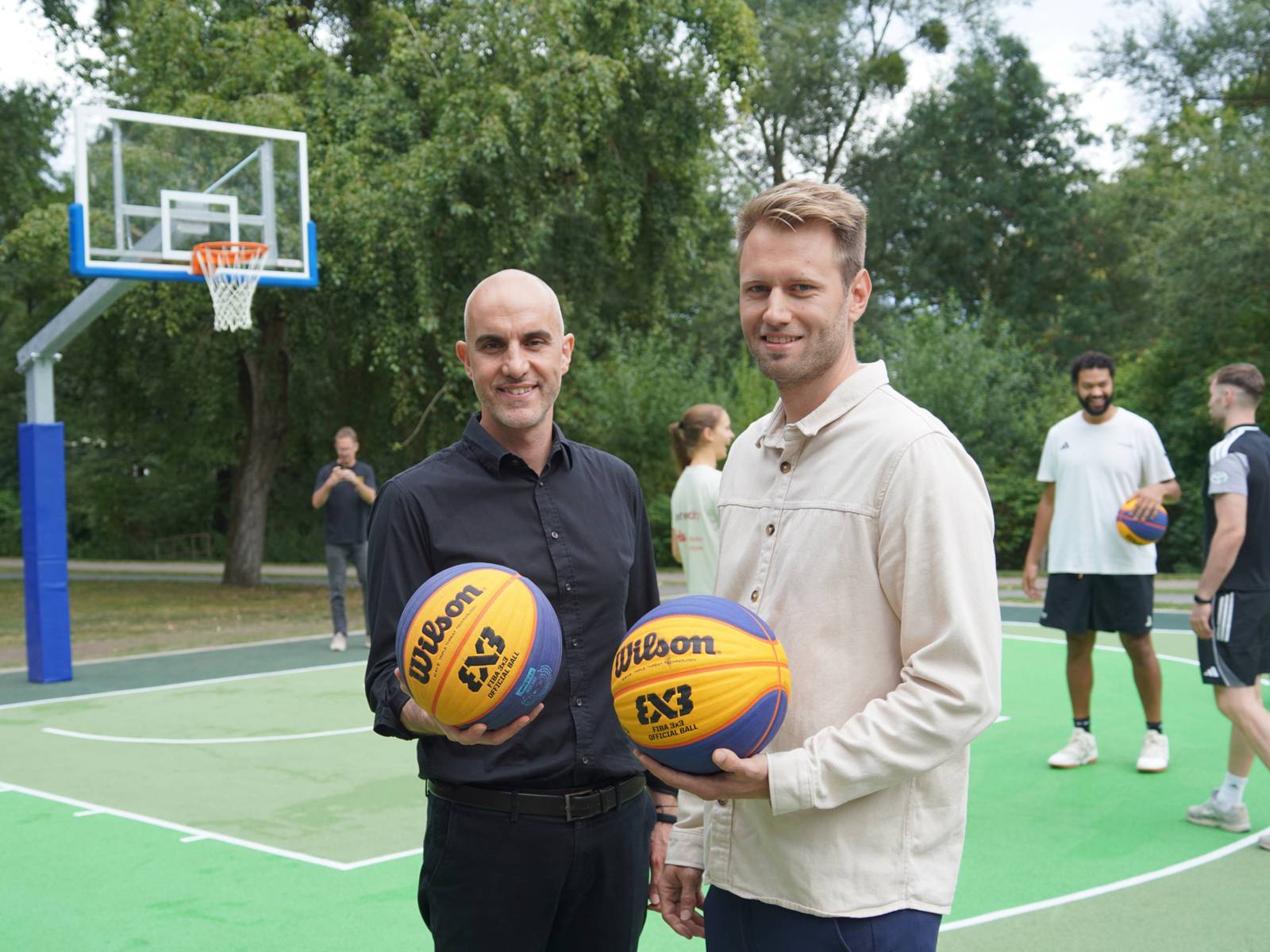 Zwei Männer stehen auf einem grünen Spielfeld und halten je einen Basketball in den Händen. Hinter ihnen ein Basketballkorb und 3 Personen in Sportkleidung.