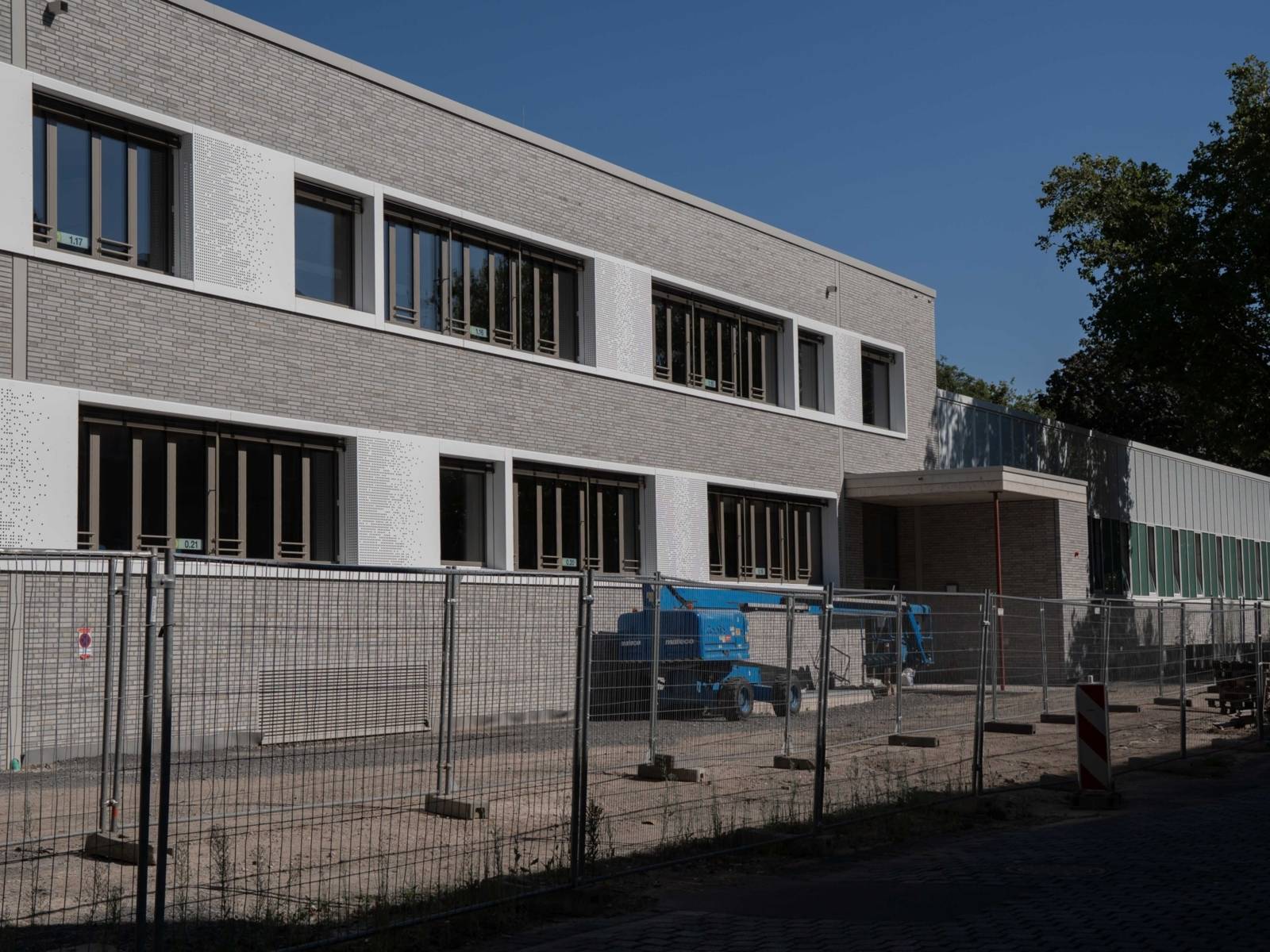 Ein Bauzaun steht vor einem zweigeschossigen Neubau mit grau-wei&szlig;er Fassade und Fensterfronten.