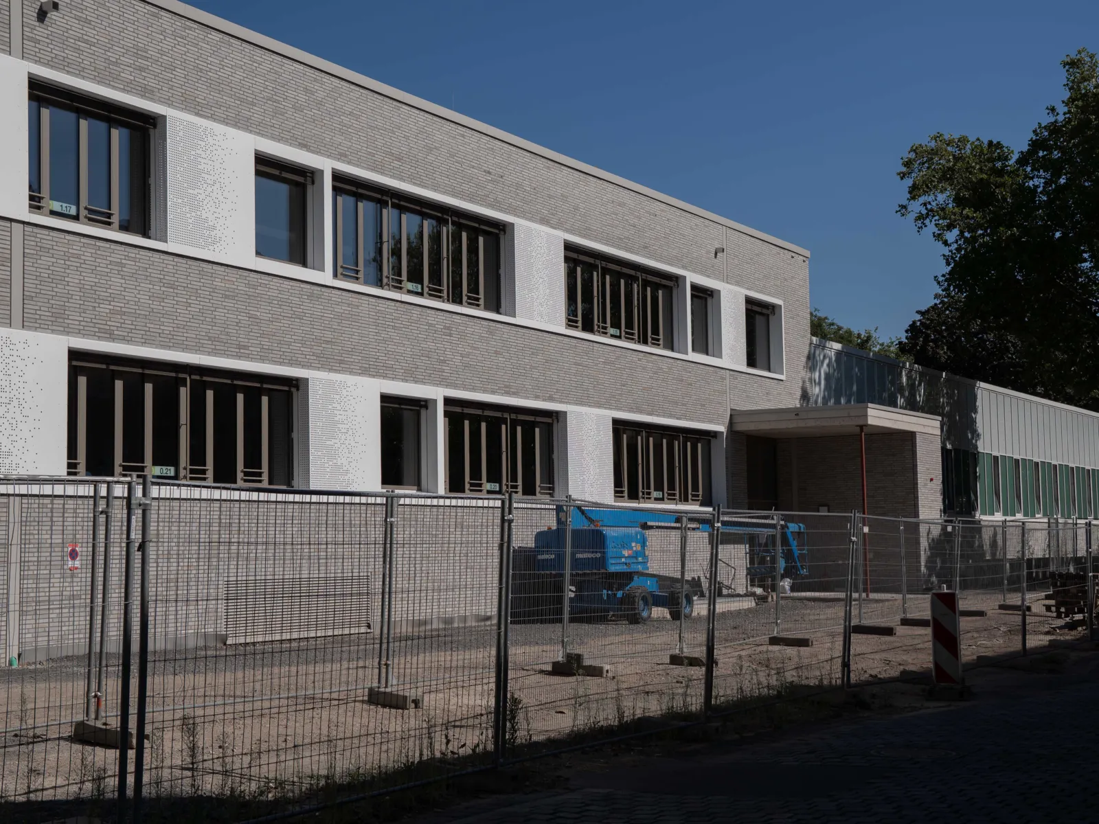 Ein Bauzaun steht vor einem zweigeschossigen Neubau mit grau-wei&szlig;er Fassade und Fensterfronten.