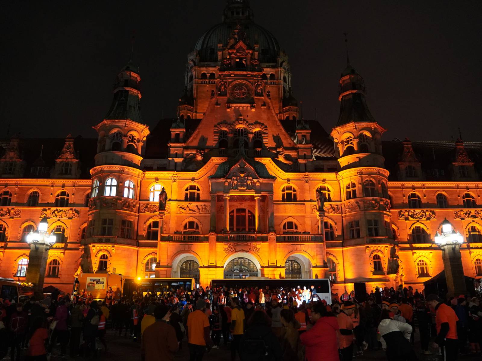 Ein altes schlossähnliches Gebäude im Dunkeln im orangem Licht angestrahlt. Davor stehen einige Menschen.