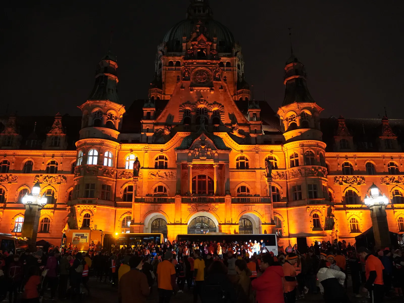 Ein altes schlossähnliches Gebäude im Dunkeln im orangem Licht angestrahlt. Davor stehen einige Menschen.