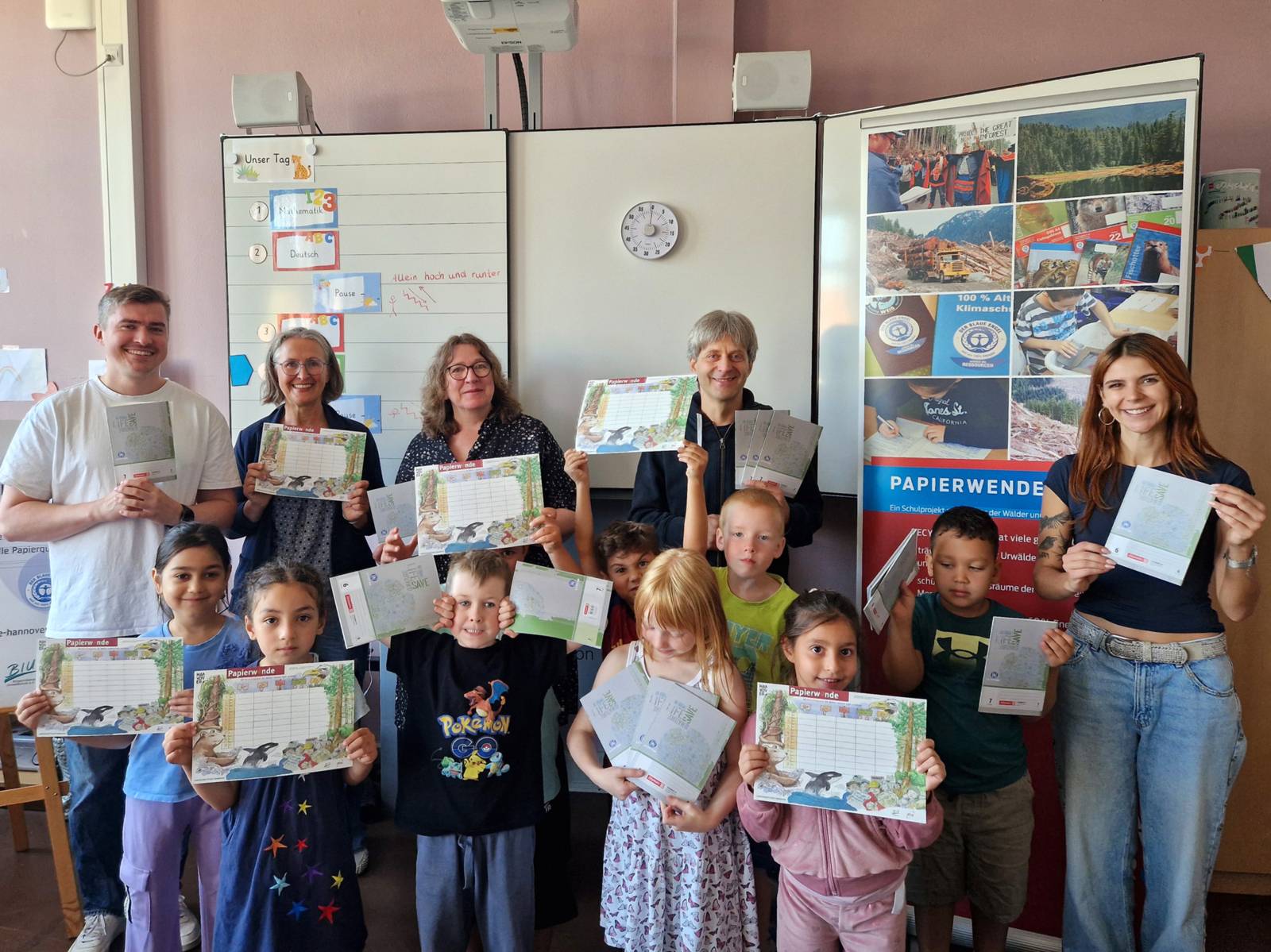 In einem Raum halten Menschen Stundenpläne und Schulhefte in den Händen und zeigen sie in die Kamera. In hinterer Reihe stehen fünf Erwachsene, vorne acht Kinder.