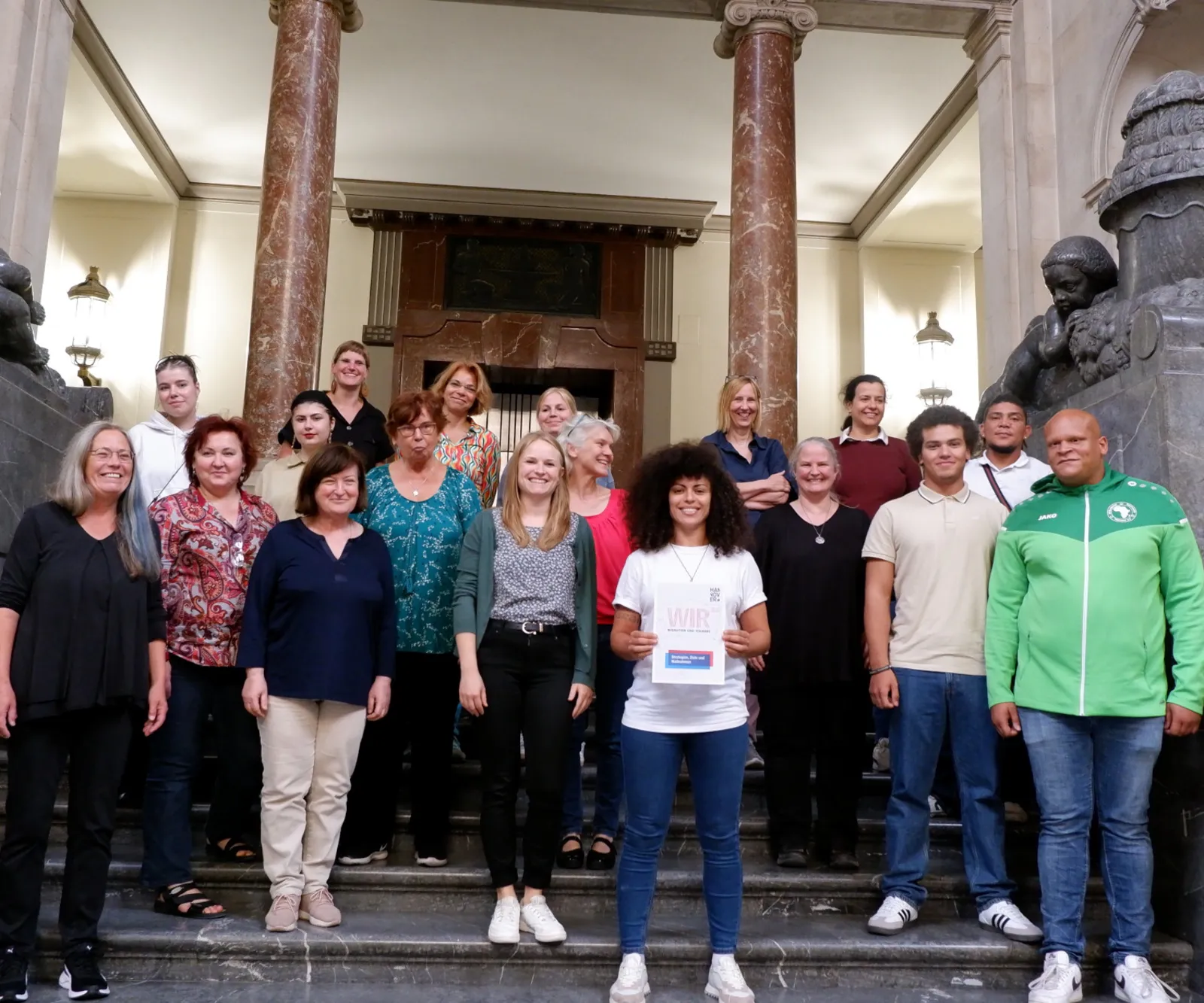 Gruppenbld: 18 Menschen, mehr Frauen als Männer, stehen auf der großen Treppe im Neuen Rathaus und lächeln in die Kamera.