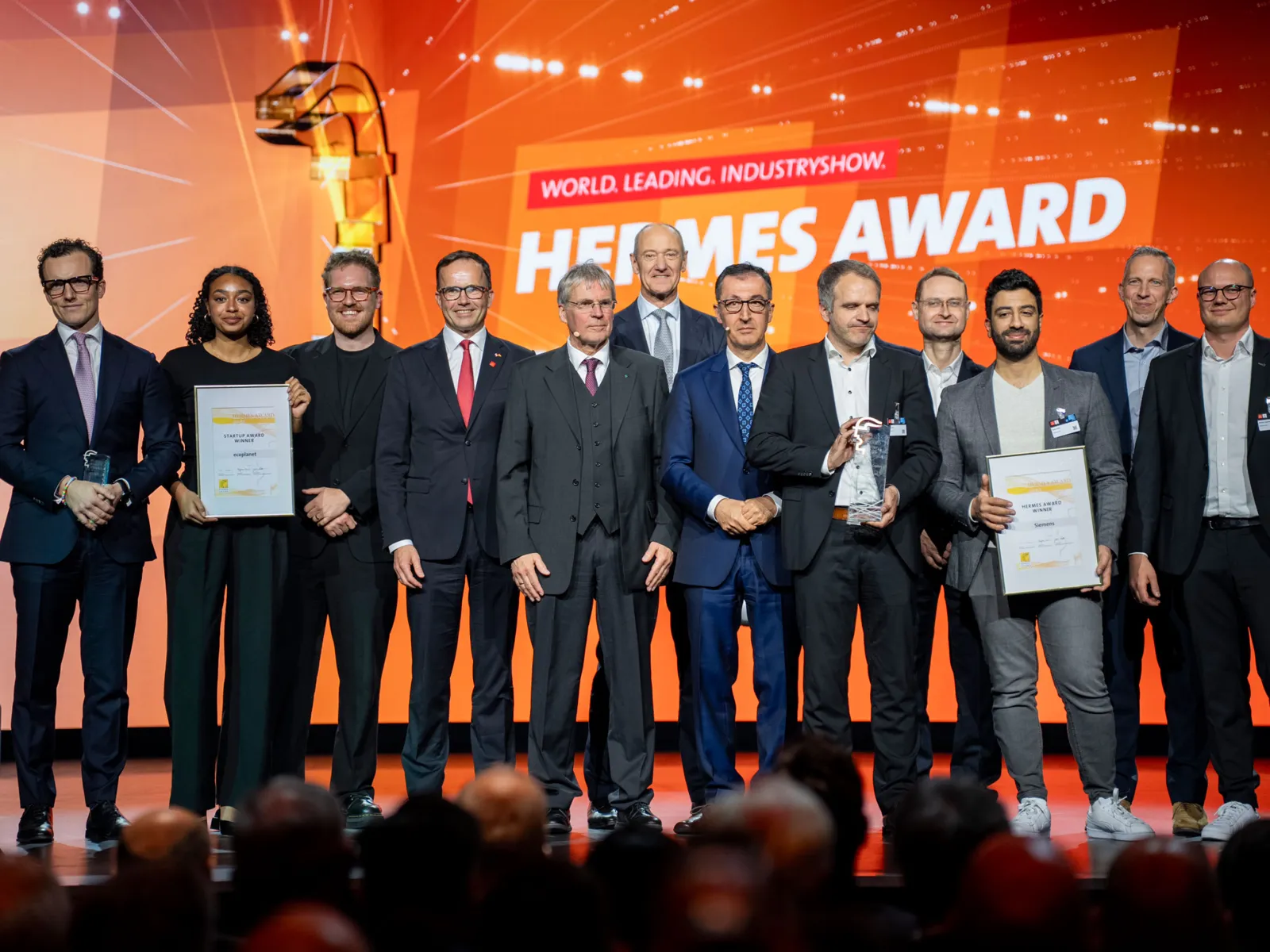 Menschen auf einer Bühne, einige halten Urkunden in den Händen, im Hintergrund der Schriftzug "Hermes Award".