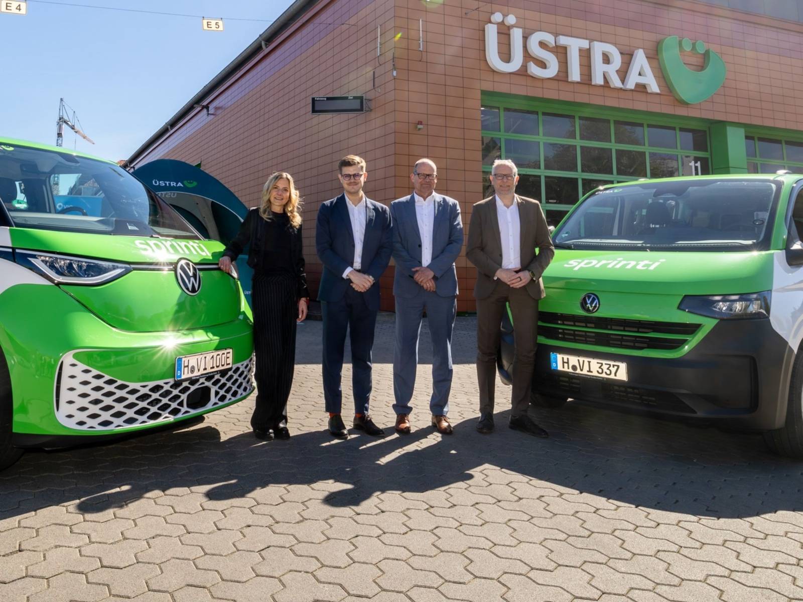 Drei Männer und eine Frau stehen nebeneinander, links und rechts daneben E-Autos von VW mit der Aufschrift "Sprinti".