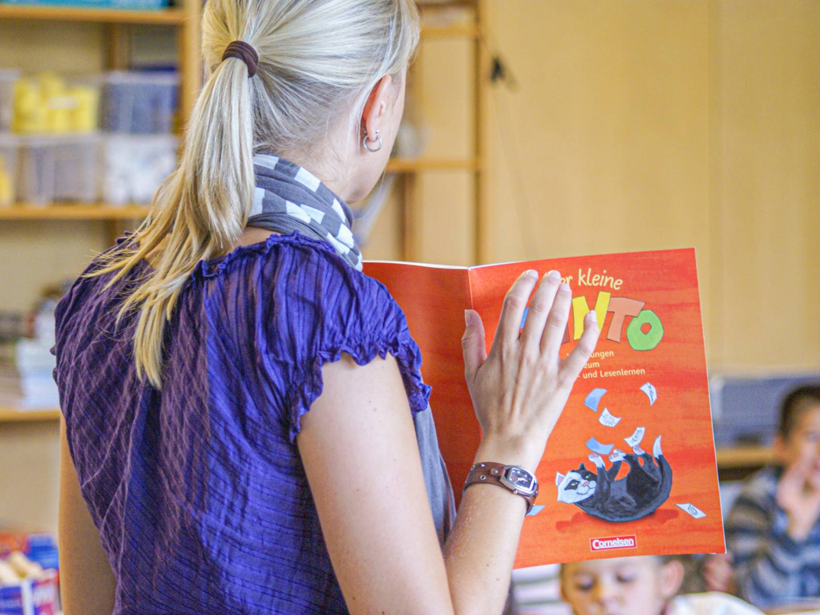 Eine Frau steht vor einer Schulklasse und hat ein Schulbuch in den Händen, man sieht den Hinterkopf und den Rücken der Frau