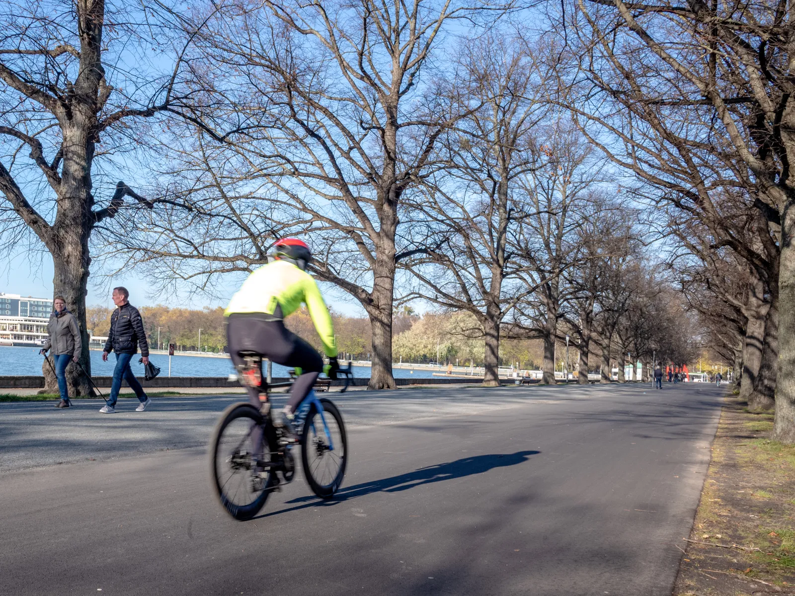 Ein Radfahrer auf einem Weg von hinten, ums&auml;umt von kahlen B&auml;umen, links daneben zwei Fu&szlig;g&auml;nger*innen und ein See.