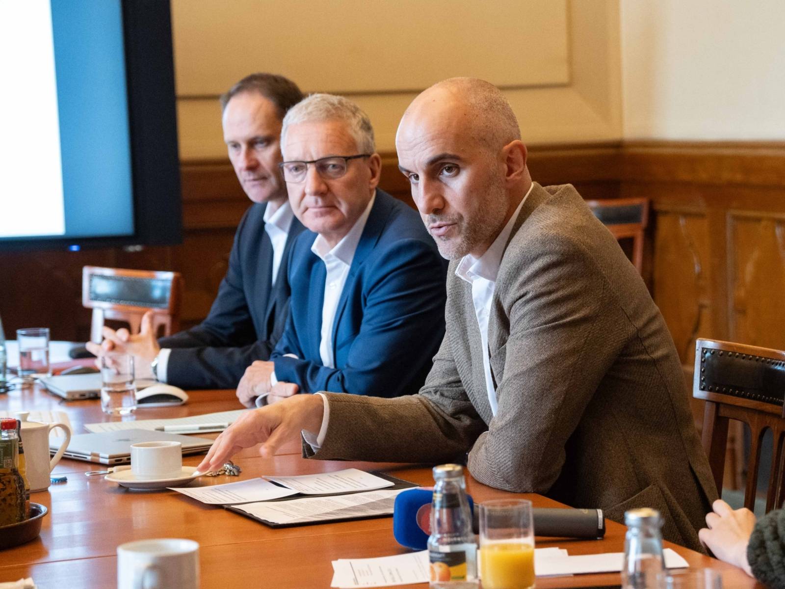 Drei Männer sitzen nebeneinander an einem Tisch, leicht seitlich dargestellt. Der vordere spricht.