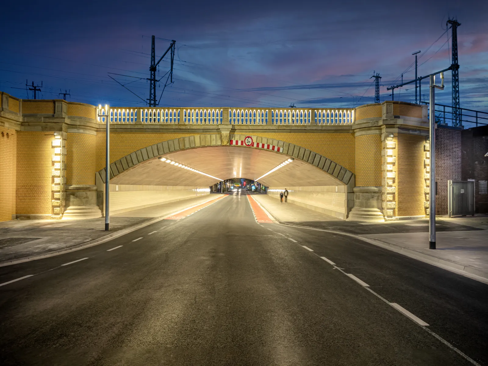 Eine Stra&szlig;e f&uuml;hrt in einen beleuchteten Tunnel, im Tunnel von Weitem zwei Personen.