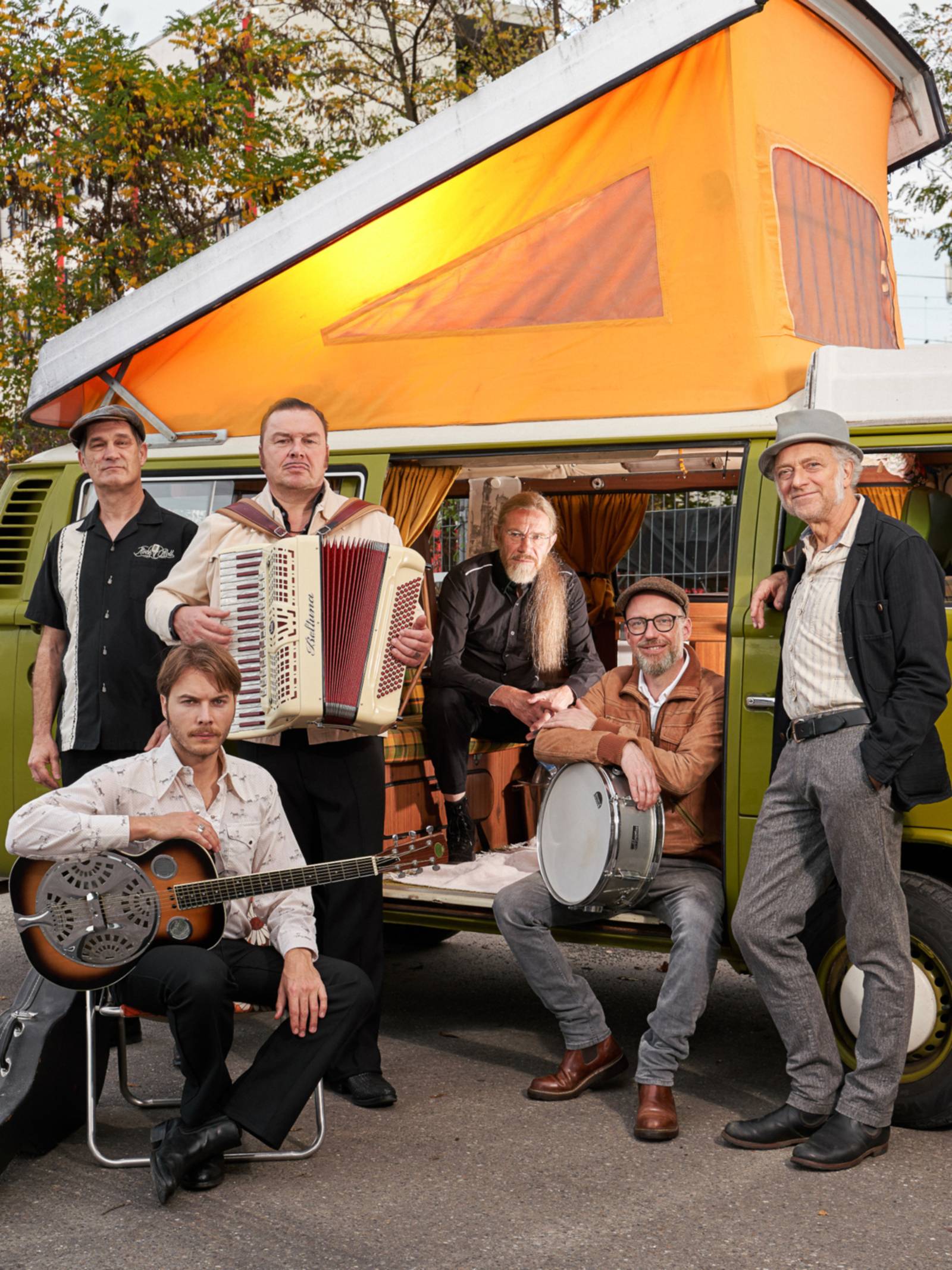 Sechs Musiker posieren mit ihren Instrumenten vor und in einem Kleinbus / Mini-Camper.