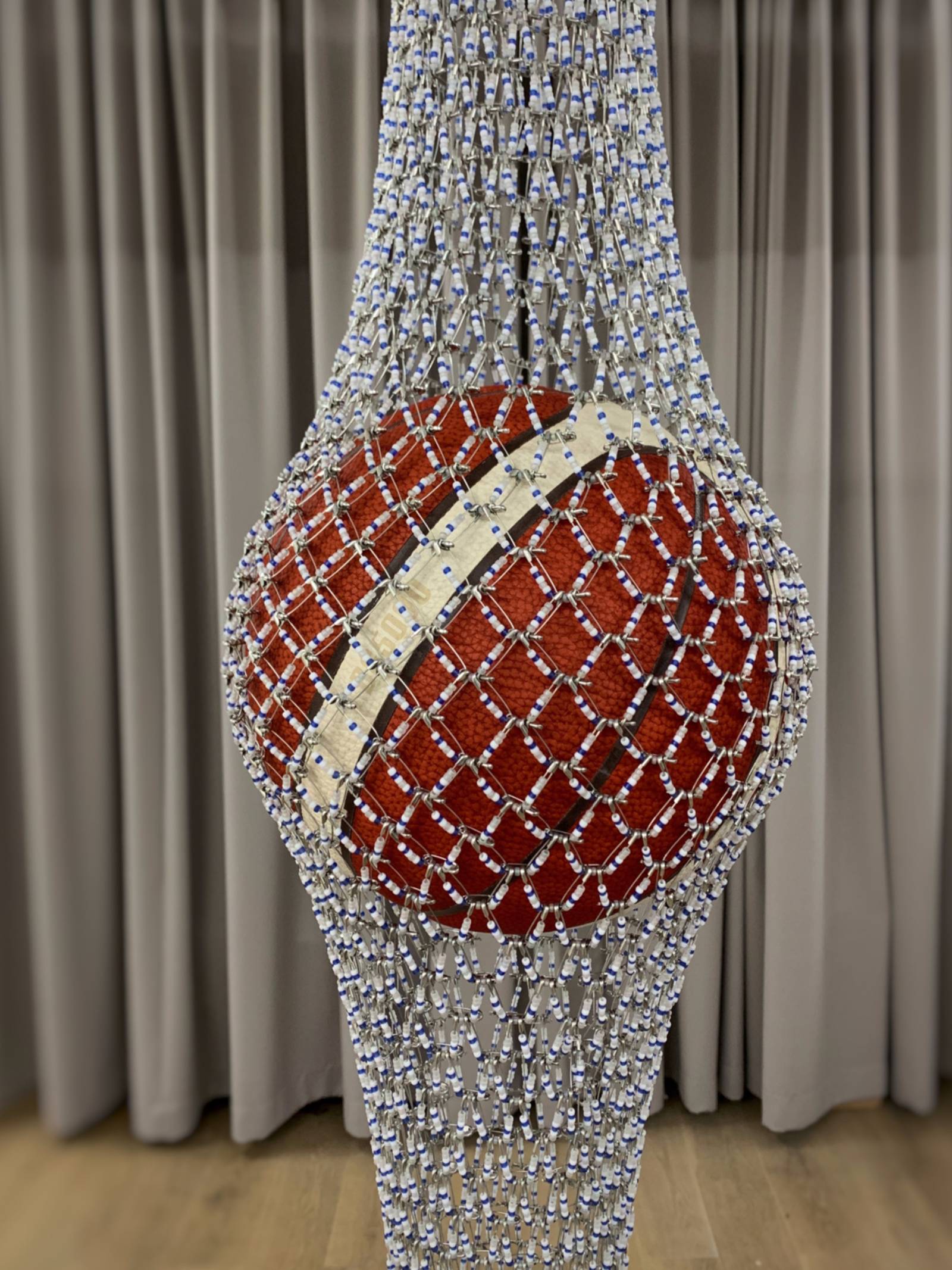 Blick auf ein Kunstwerk von Shige Fujishiro: Ein Basketball wird von einem dünnen Netz gehalten, auf den Fäden des Netzes sind Ziersteine. Im Hintergrund ist ein Vorhang.