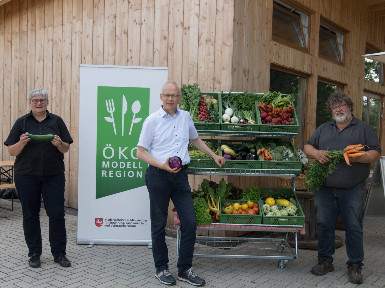 Drei Personen, die Gemüse in den Händen halten, stehen vor einem Schild der Ökomodellregion und einem Gemüsestand