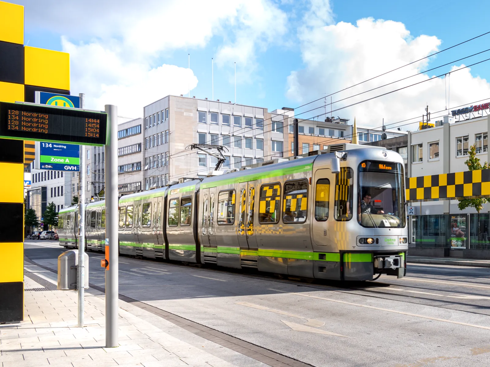Stra&szlig;e, eine Haltstelle und eine Stadtbahn, dahinter Geb&auml;ude