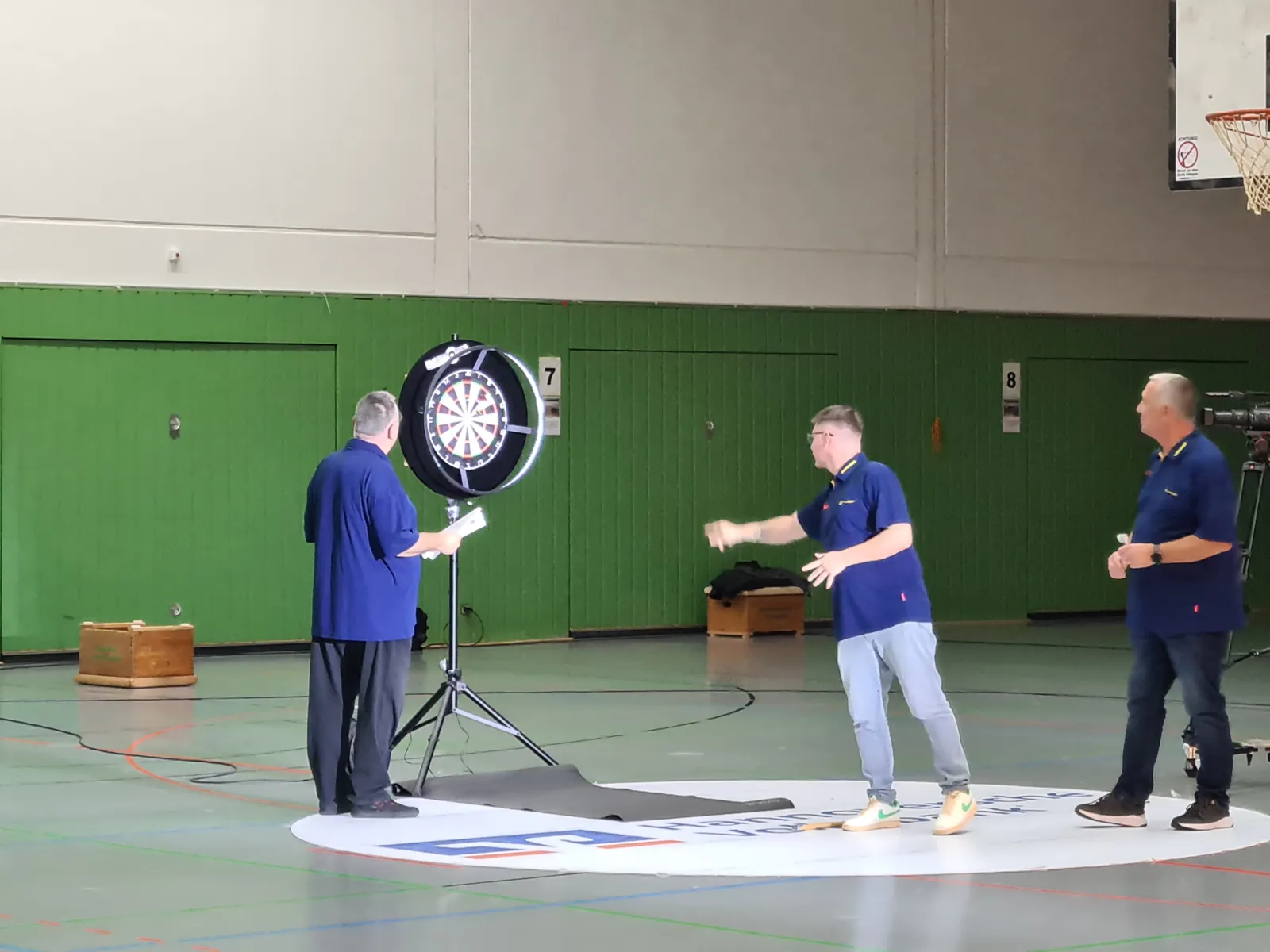 Auf dem Bild sind drei M&auml;nner zu sehen, die in einer Turnhalle Dart spielen.