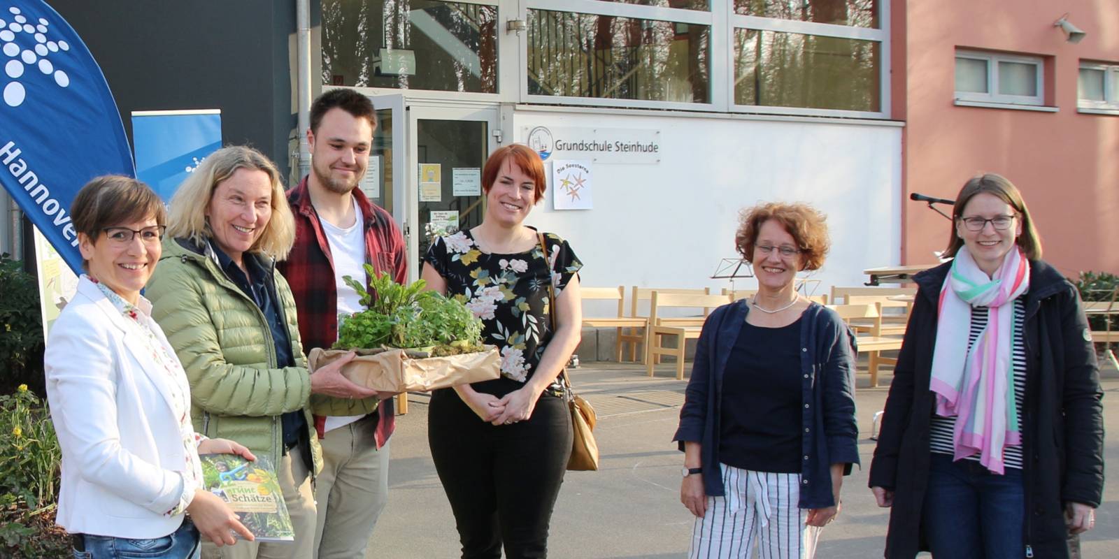 Fünf Frauen und ein Mann stehen vor der Grundschule Steinhude. Eine Frau gibt einem Mann eine Kiste mit Pflanzen.