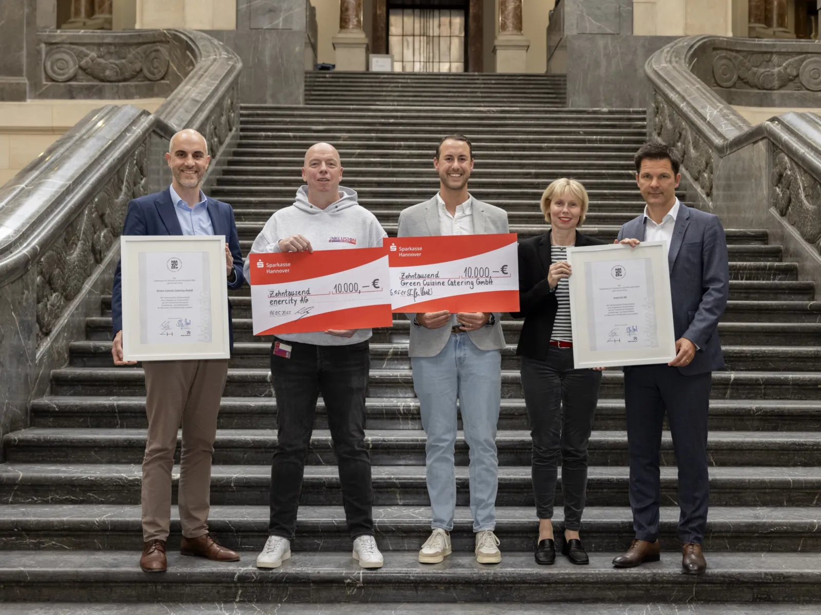 Fünf Personen stehen auf einer Treppe und halten zwei Urkunden und zwei überdimensionale Schecks in den Händen.