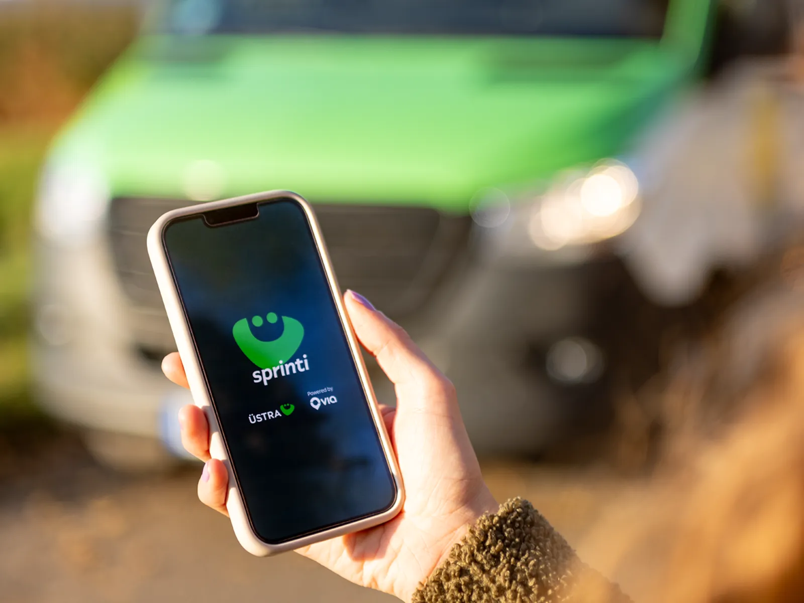 Eine Frau steht im Freien an einer Straße und schaut auf ihr Handy. Ein Transporter nähert sich.