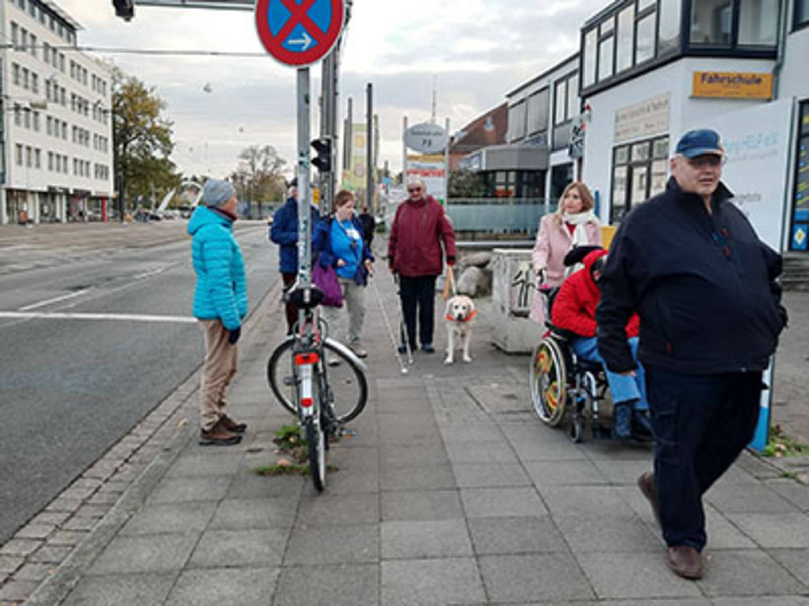 Menschen und Menschen mit Behinderung (Blinde Personen, eine Person mit Blindenhund und eine Person im Rollstuhl) auf der Sutelstraße.