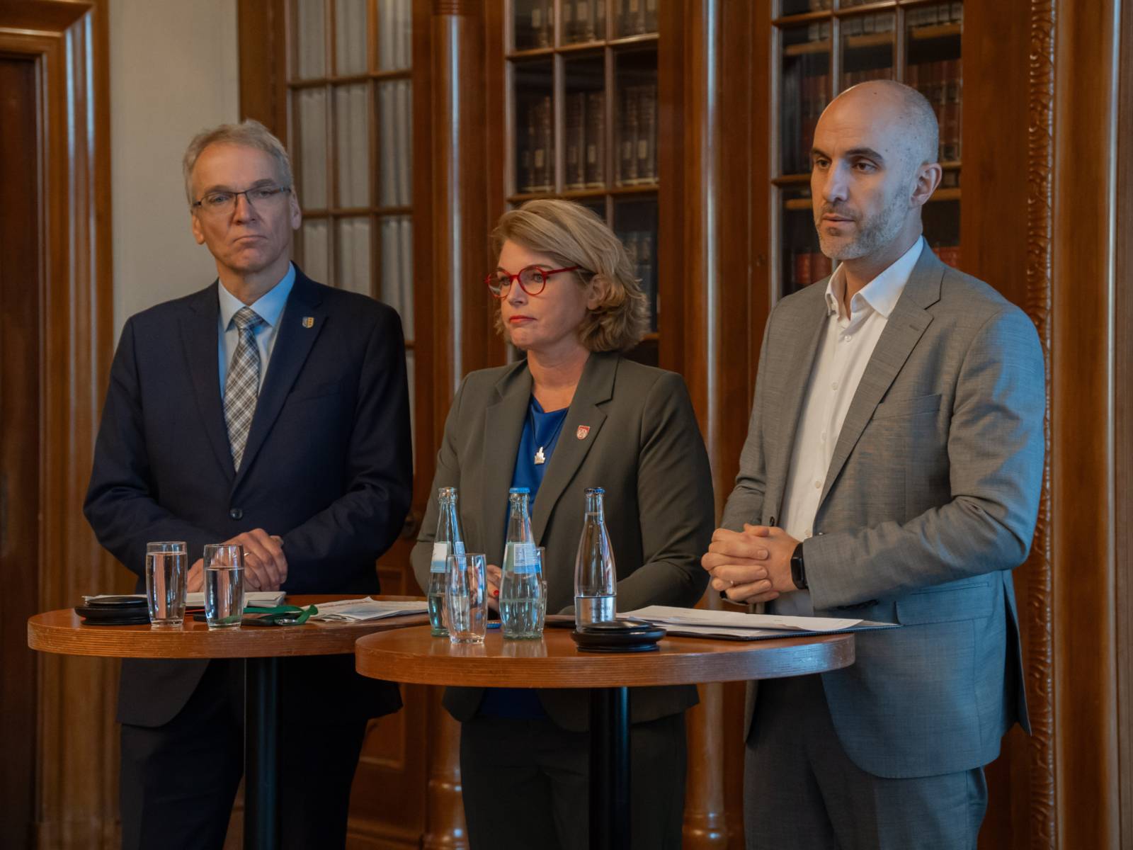 Drei Personen stehen an Stehtischen, vor ihnen stehen Wassergläser.