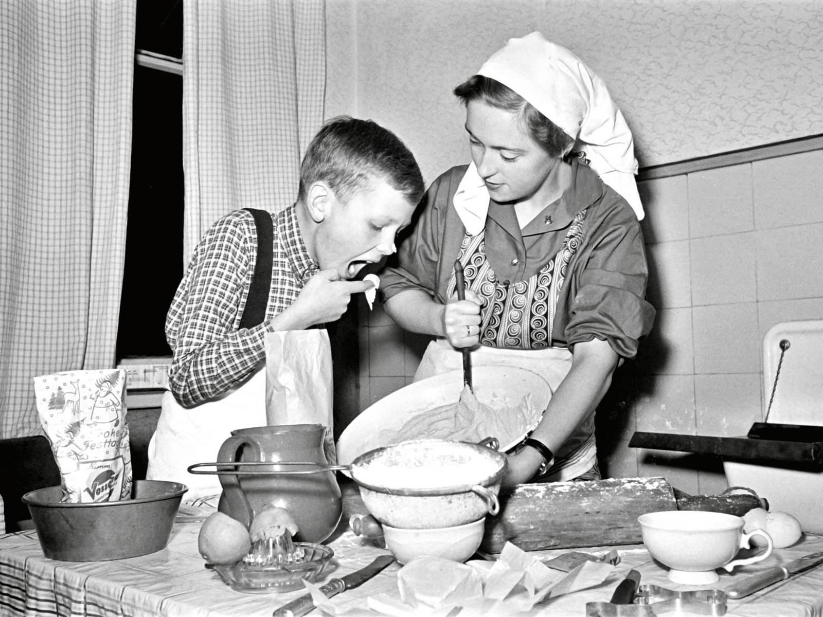 Schwarz-Weiß-Fotografie: Eine Frau trägt Kopftuch und Schürze und rührt mit der Hand einen Kuchenteig. Neben ihr ist ein Junge, der mit dem Finger Teig probiert, der Junge hat auch Kuchenteig an der Nasenspitze.