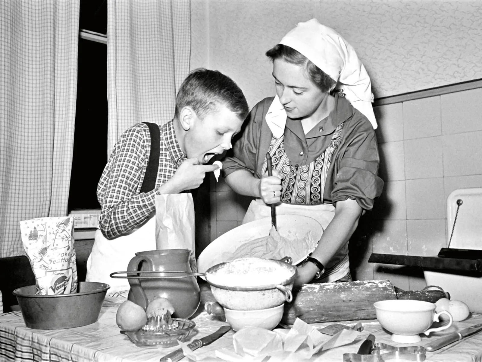 Schwarz-Wei&szlig;-Fotografie: Eine Frau tr&auml;gt Kopftuch und Sch&uuml;rze und r&uuml;hrt mit der Hand einen Kuchenteig. Neben ihr ist ein Junge, der mit dem Finger Teig probiert, der Junge hat auch Kuchenteig an der Nasenspitze.