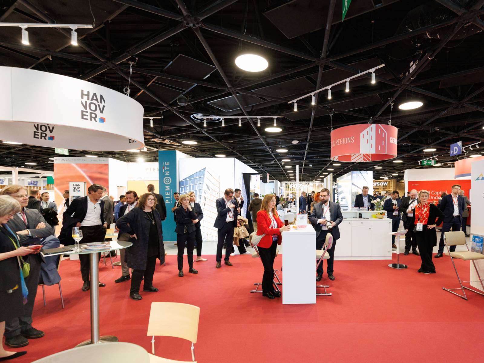 Menschen stehen in einer Halle mit Stehtischen und Ständen.